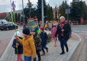 Dzieci z nauczycielkami idą po chodniku niosąc kolorowe ozdoby