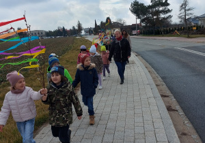 Dzieci z nauczycielkami idą po chodniku niosąc kolorowe ozdoby