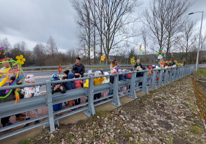 Dzieci z nauczycielkami stoją na moście trzymając kolorowe dekoracje