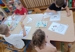 Dzieci siedzą przy stoliku i wypełniają farbą kropeczki