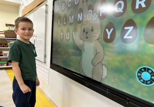 04 Uśmiechnięty chłopiec stoi przed tablicą interaktywną, na któej widać jajeczka z alfabetem i króliczka.