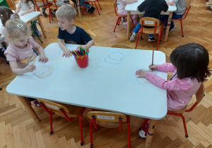Dzieci siedzą przy stolikach i rysują emocje na przygotowanych szablonach twarzy.