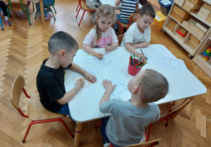 Dzieci siedzą przy stolikach i rysują emocje na przygotowanych szablonach twarzy.