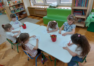 Dzieci siedzą przy stolikach i rysują emocje na przygotowanych szablonach twarzy.