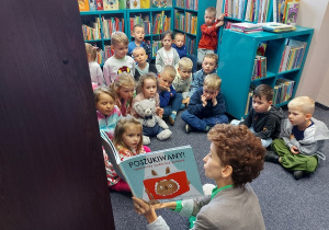 Przedszkolaki siedzą na dywanie i słuchają książki czytanej przez bibliotekarkę.