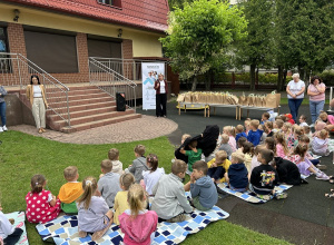 Pani Dyrektor stojąca przy stolikach z nagrodami przemawia do dzieci siedzących na kocach