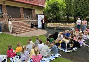 05 Pani Dyrektor stojąca przy stolikach z nagrodami przemawia do dzieci siedzących na kocach