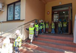 Dzieci ubrane w odblaskowe kamizelki z kijami w dłoni wychodzą z przedszkola.