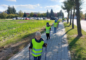 Przedszkolaki z kijami w dłoni maszerują chodnikiem.