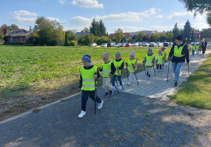 Skupione przedszkolaki z kijami w dłoni maszerują chodnikiem.