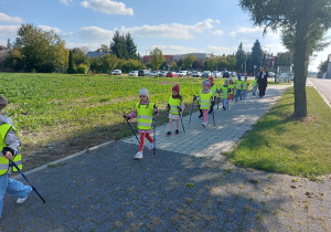Zadowolone przedszkolaki z kijami w dłoni maszerują chodnikiem.
