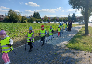 Przedszkolaki z kijami w dłoni maszerują chodnikiem.