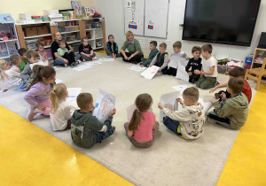 Dzieci siedzą w kole na dywanie ze swoimi pracami.