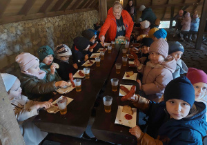 Dzieci siedząc przy stole jedzą kiełbaski.