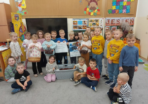 Przedszkolaki trzymające plakaty informujące o zbiórce, ustawione przy koszu z kjasztanami.