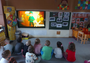 Przedszkolaki siedzą przed ekranem i oglądają bajkę edukacyjną.