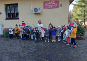 Dzieci z kijkami i biało-czerwonymi flagami stoją na tle budynku przedszkola