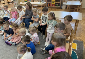 Dzieci siedza na dywanie i słuchaja prowadzącego.