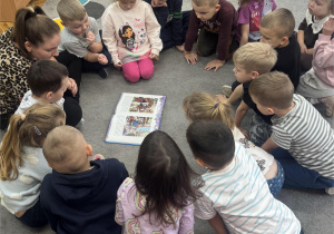 Dzieci siedzą w kole i oglądają ilustracje w książce.