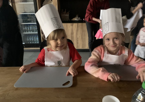 Dziewczynki siedzą przy stole ubrane w fartuszki.