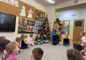 Chłopiec siedzi na kolanach swojej mamy a dzieci siedzą w kole i słuchają bajki.