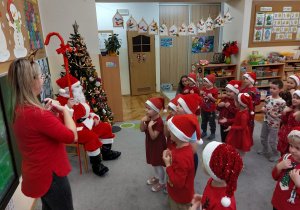 Dzieci ubrane w mikołajkowe czapeczki i czerwone stroje tańczą dla mikołaja