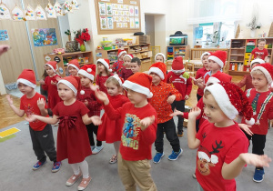 Dzieci ubrane w mikołajkowe czapeczki i czerwone stroje tańczą dla mikołaja