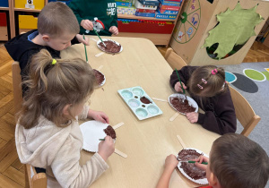 Dzieci siedzą przy stoliku i malują papierowe talerzyki.