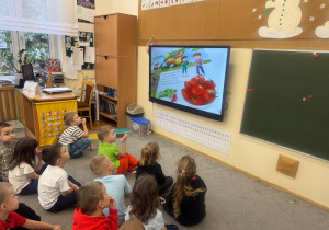 Dzieci siedzą na dywanie i słuchają informacji zawartych w filmie edukacyjnym.