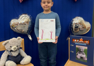 Chłopiec prezentuje swoją pracę w ramach projektu „Mały Miś w Świecie Wielkiej Literatury” – obok siedzi Miś, a na drugim krześle znajduje się Dzienniczek Małego Misia.