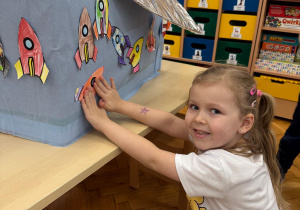 Dziewczyka z dumą prezentuje i przykleja swoją pracę.