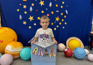 Chłopiec siedzi w statku kosmicznym ozdobionym rakietami i pozuję na tle gwiazd i planet.