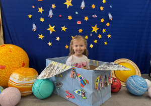 Dziewczynka bawi się w astronautę, siedząc w dekorowanym statku z rakietami.