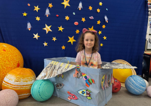 Dziewczynka uśmiecha się na tle gwiazd i planet, siedząc w kosmicznym pojeździe z pudełka.