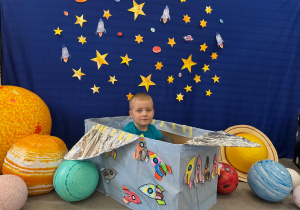 Chłopiec pozuję do zdjęcia na tle kosmicznej dekoracji z rakietami i planetami.