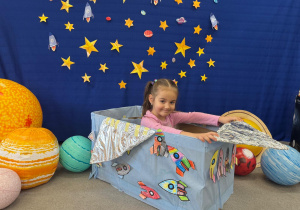 Dziewczynka uśmiecha się na tle gwiazd i planet, siedząc w kosmicznym pojeździe z pudełka.