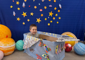 Chłopiec siedzi w statku kosmicznym ozdobionym rakietami i pozuję na tle gwiazd i planet.