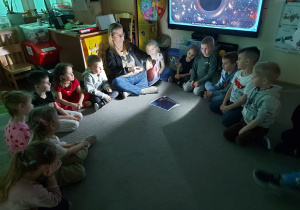 Dzieci siedzą w kole i oglądają obrazki dotyczące kosmosu podświetlone latarką.