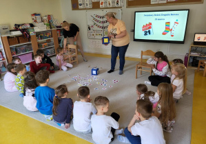 Dzieci siedzą na dywanie i słuchają nauczyciela.