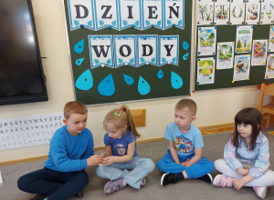 Przedszkolaki w niebieskich strojach podają kubek z wodą.