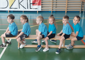 Grupka dzieci siedzi na ławce, czekając na polecenie nauczyciela.