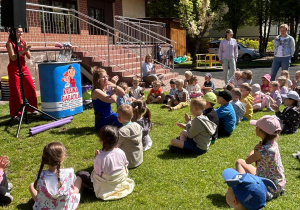Dzieci siedzą na trawie i wykonują ruchy rąk do zabawy.